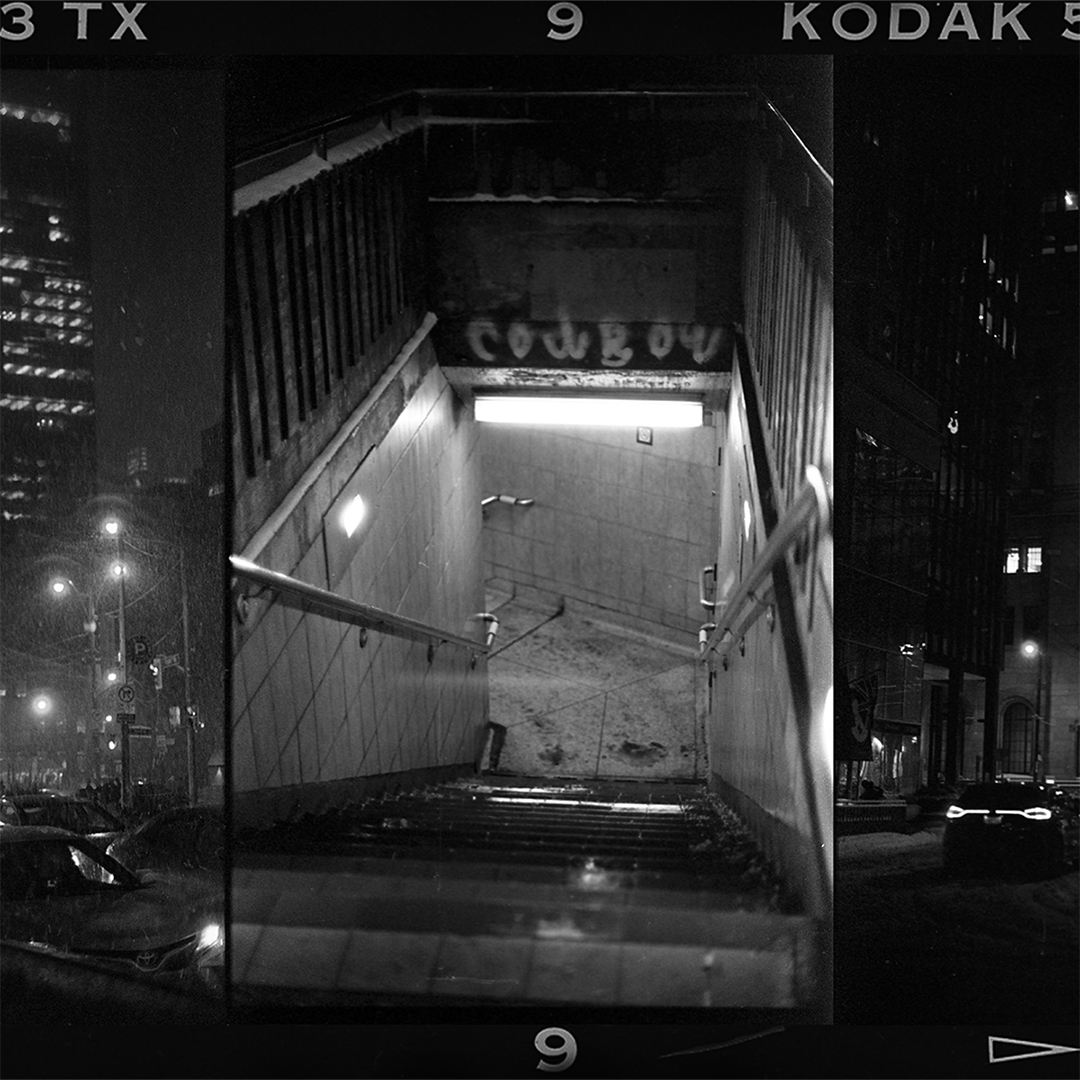 Looking down the stairs of a subway staion from street level, with a narrow section of a city street at night on either side