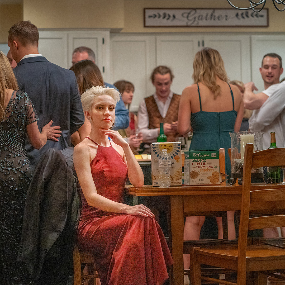 A blonde woman is sitting at a tabloe during a party with people standing around her in various unrelated conversations