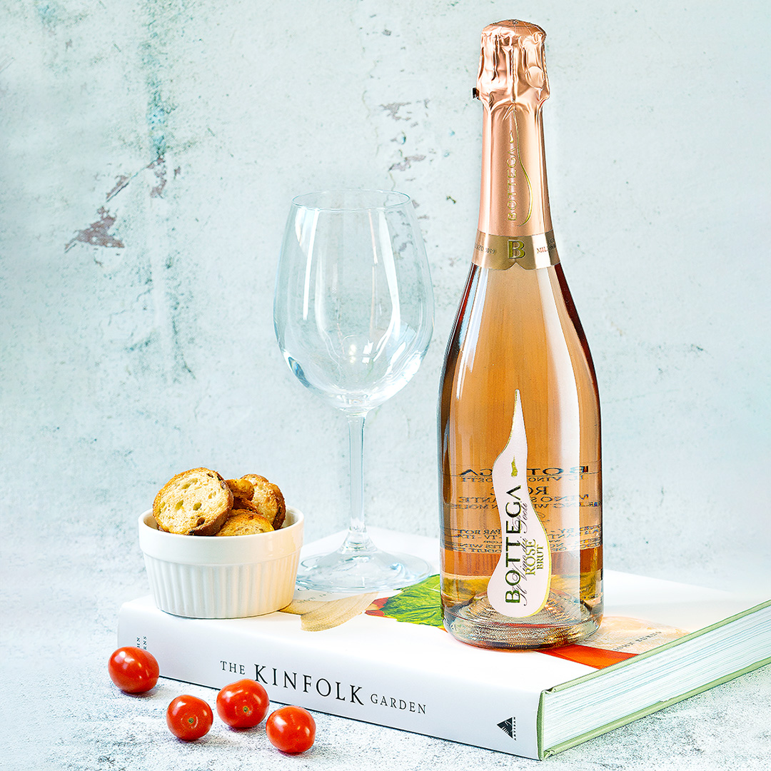 A rose bottle of sparkling wine sitting on a closed cook book, beside an empty wine glass and a dish of crackers. There are four mini tomatoes scattered in front