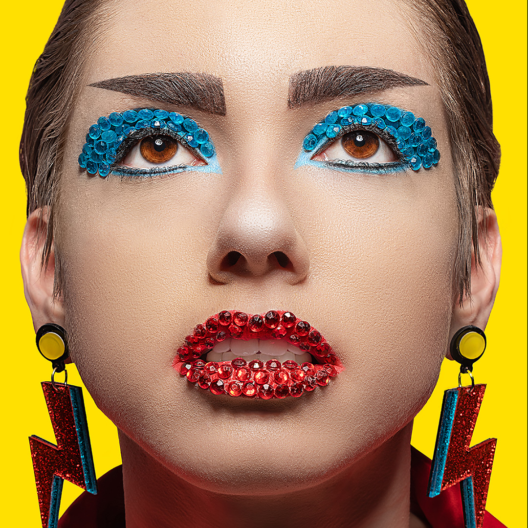 Close up head shot of a young woman with blue and red rhinestones and glitter used as makeup on eyes and lips, with a yellow backdrop.