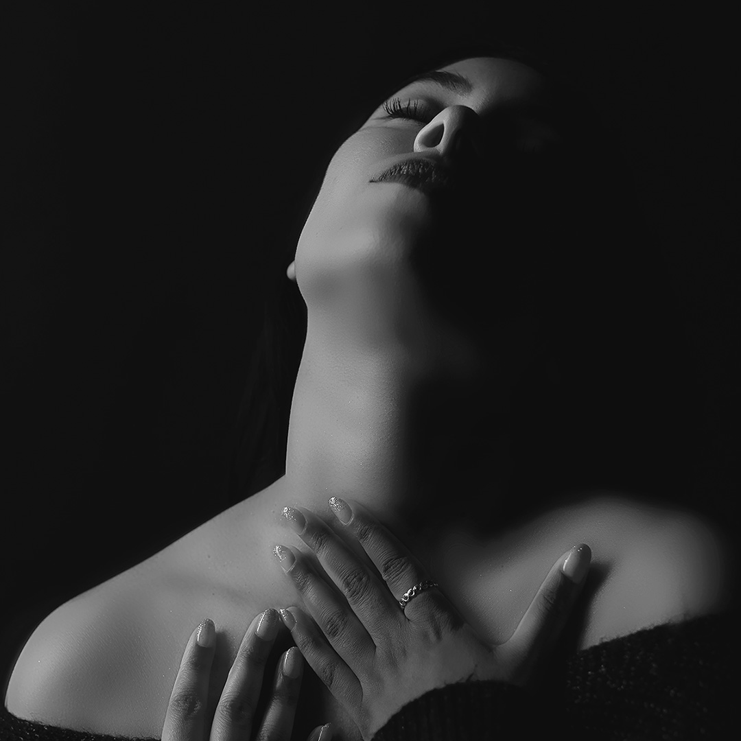 black and white image of a woman wearing a knitted sweater in a dark room with dim lighting, both her hands are softly touching her check and neck