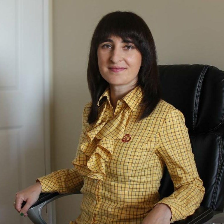 This is a headshot image of Elizaveta Dolgova. She is sitting in the office chair. She wears a yellow business casual shirt.