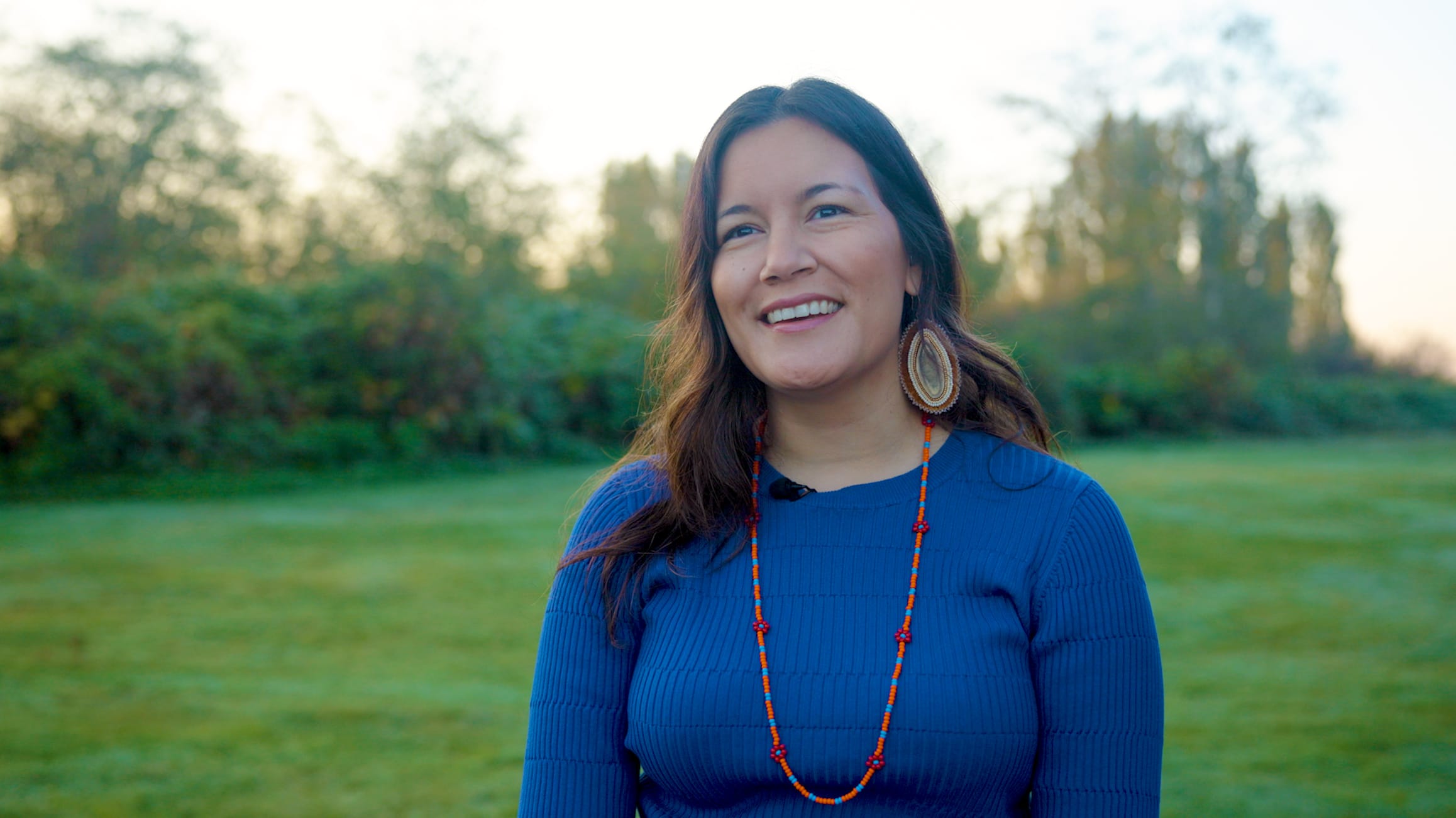 Deputy director of the Indigenous Leadership Initiative Dahti Tsetso wears a blue shirt and orange necklace in a field of grass