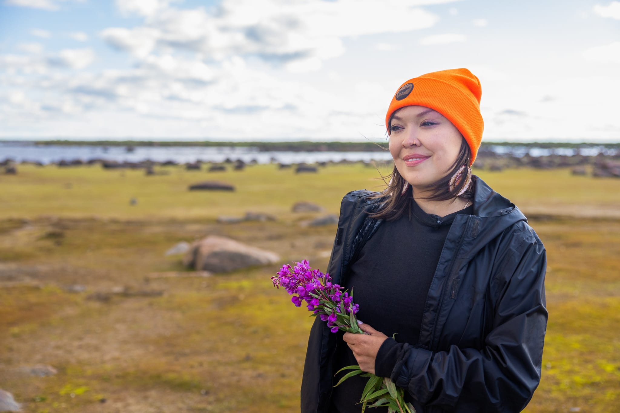 Seal River Watershed Alliance director Stephanie Thorassie holds a bundle of purple flowers while wearing an orange hat and black coat in a field of grass