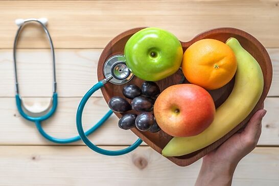 A heart shaped fruit basket with two apples, an orange, a banana, and grapes next to a stethoscope.
