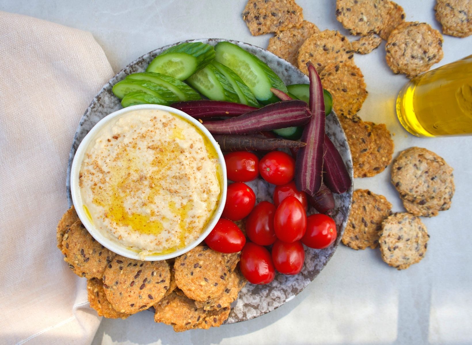Overhead shot of a white bowl of hummus, a low-glycemic snack. A plate with sliced cucumbers, cherry tomatoes, purple carrots, and whole grain crackers are on the side
