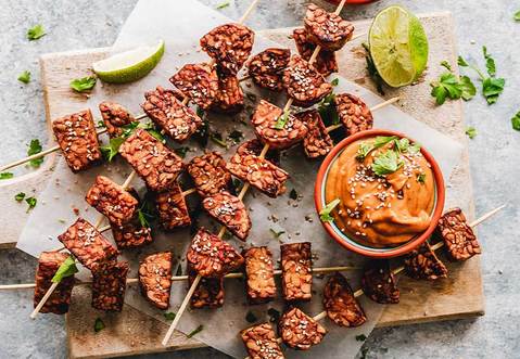 Placed on a marble table is a cutting board that has cooked tempeh skewers on it. Next to the skewers is a small bowl with sauce and some lime pieces.