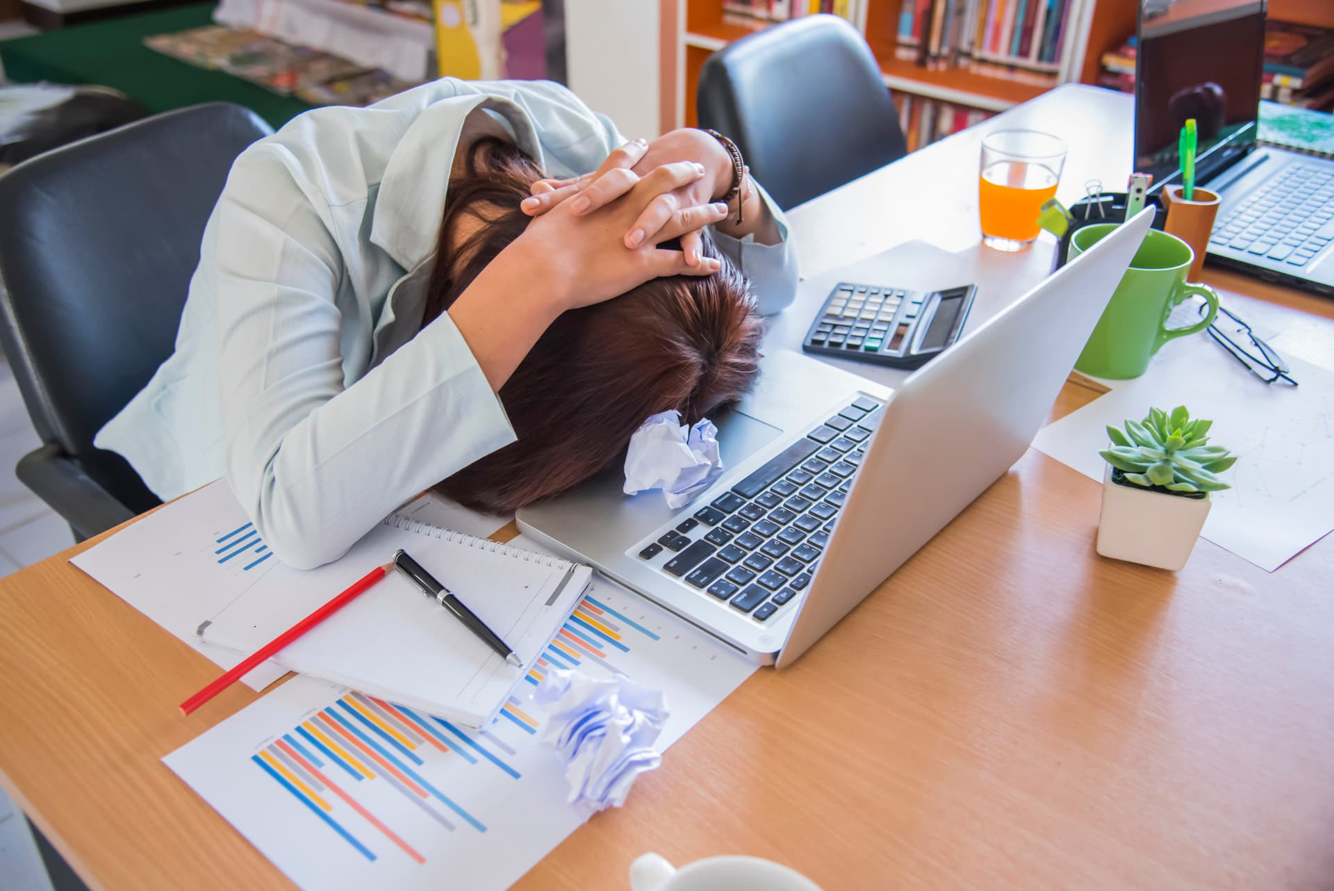 A frustrated person with their head on the desk, working on paperwork on their computer