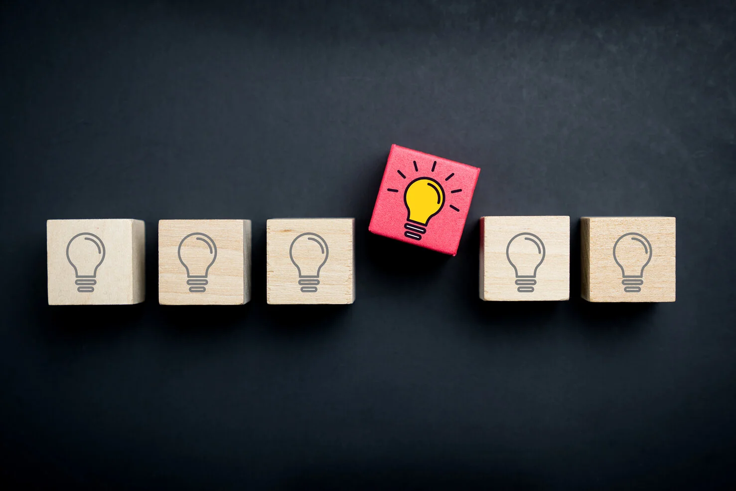 A row of wood blocks with icons of a lightbulb on each one. One in the middle is in full color, light bulb is bright yellow indicating an idea. The background is a dark chalkboard.