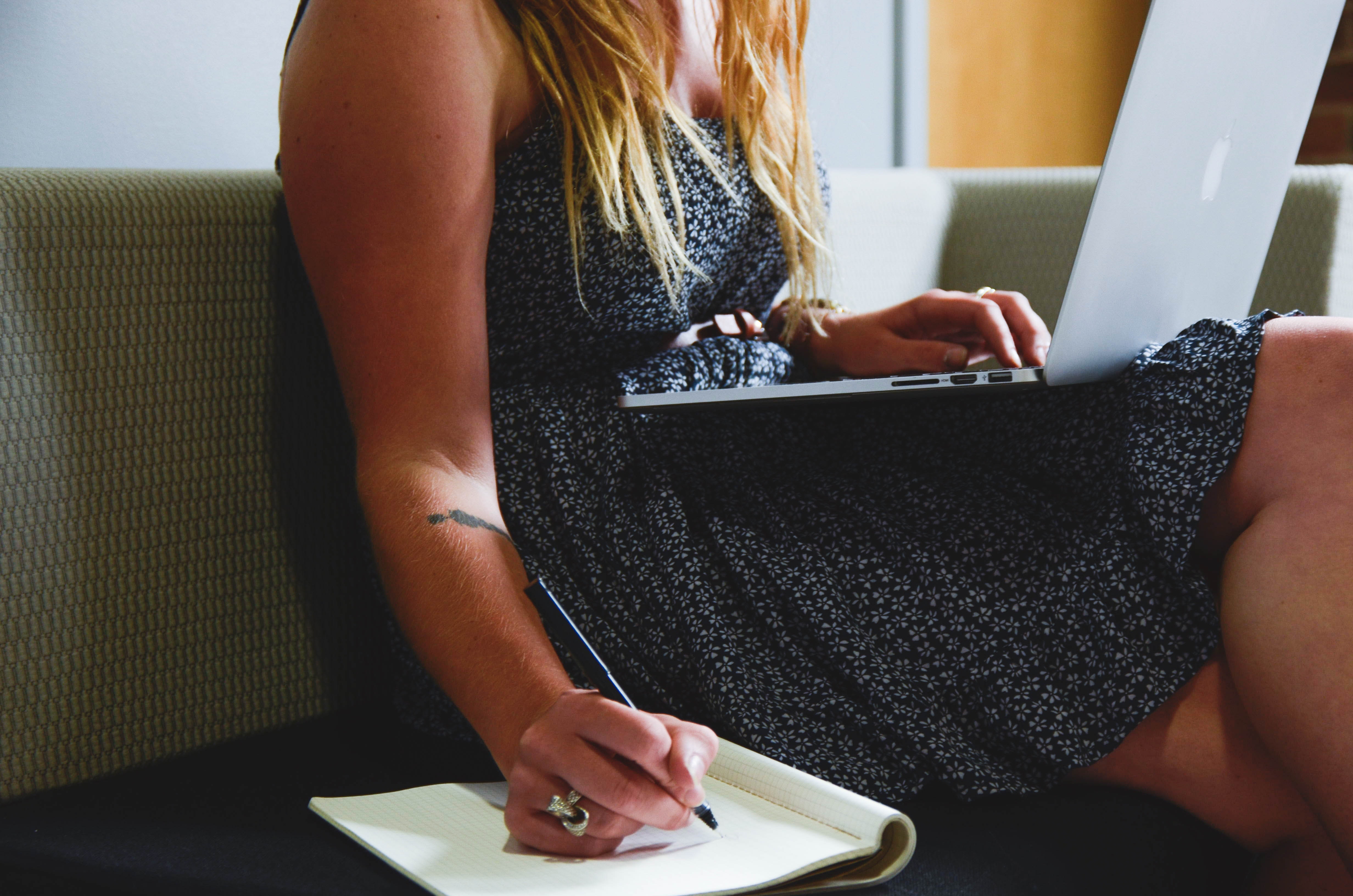 A person in a dress sitting on a couch with a laptop on their lap, writing in a notebook sitting on the couch beside them.