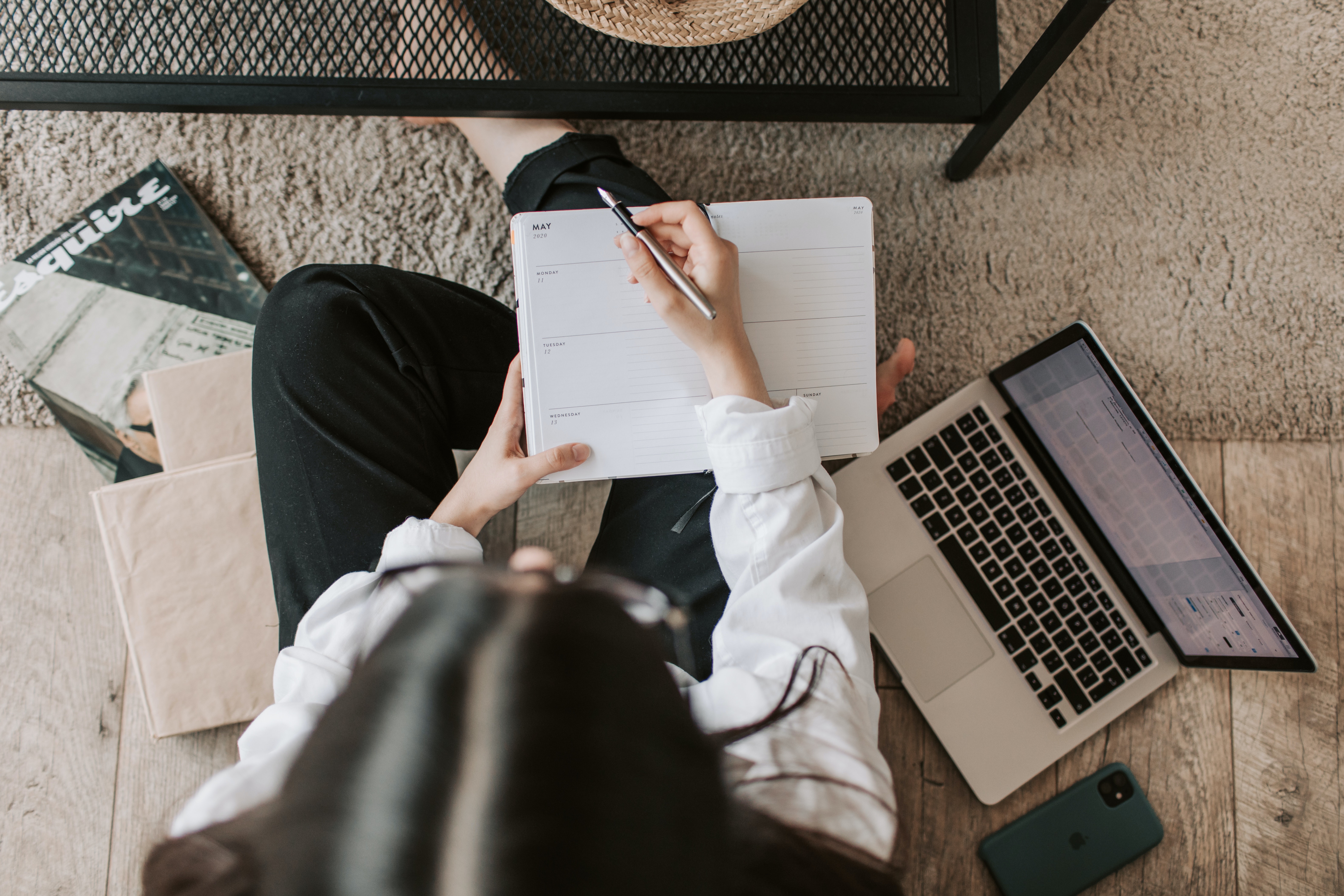 Overhead view of a person with long, dark hair sitting on a beige carpet and writing in a planner beside an open laptop.