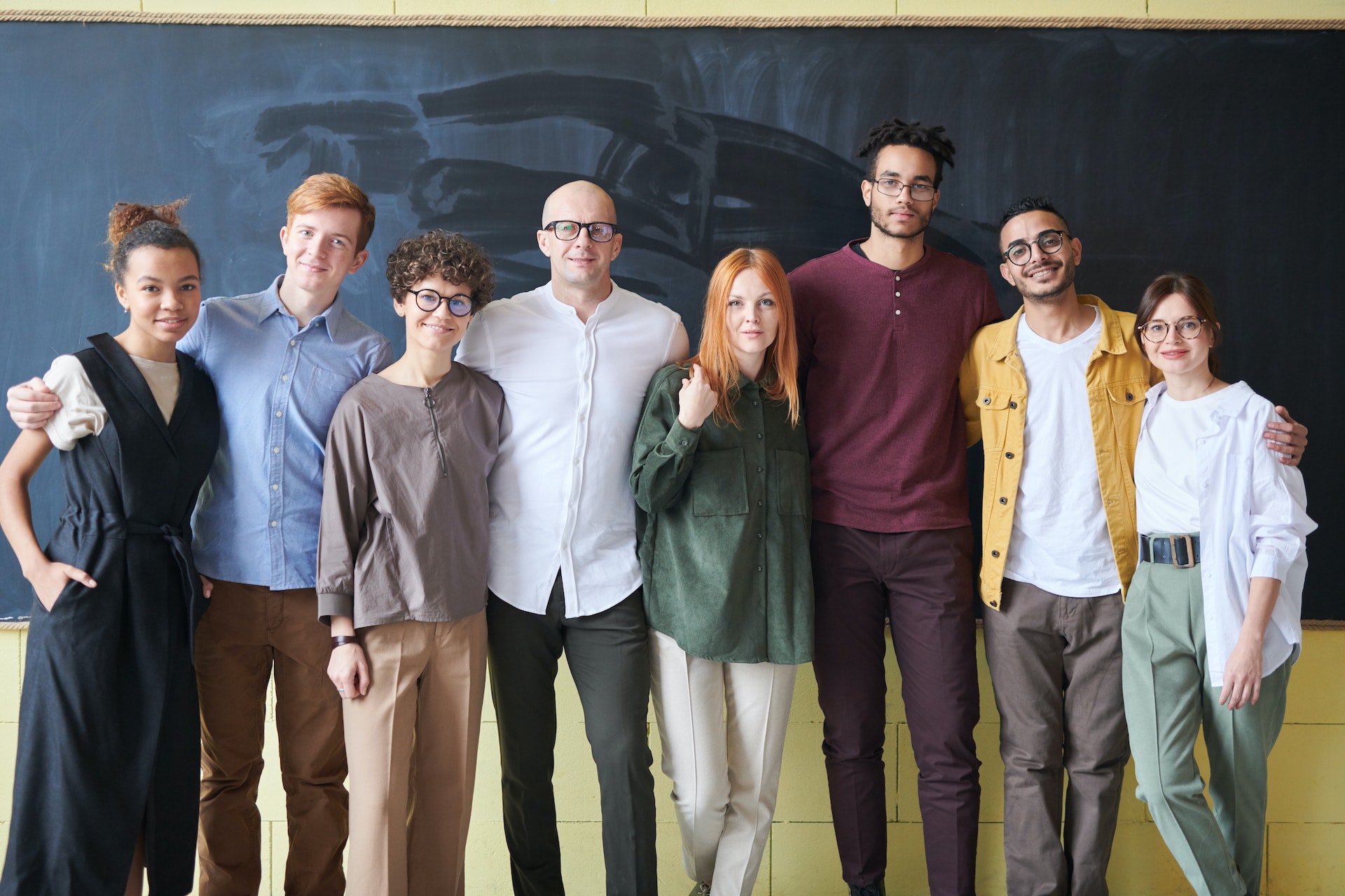 A group of smiling people stand in a line in front of a blackboard.