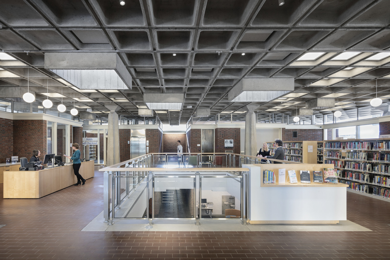 A library opening with cement and lighting panels in the ceiling, people standing around the welcome desk and bookshelves, overlooking the lower level.