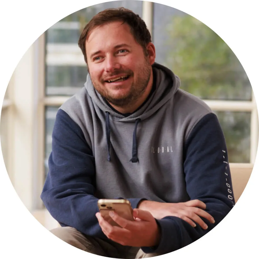 Jake sits and smiles directly at the camera in front of large windows, holding their phone. They have short brown hair and a short beard. They are wearing a blue hoodie.