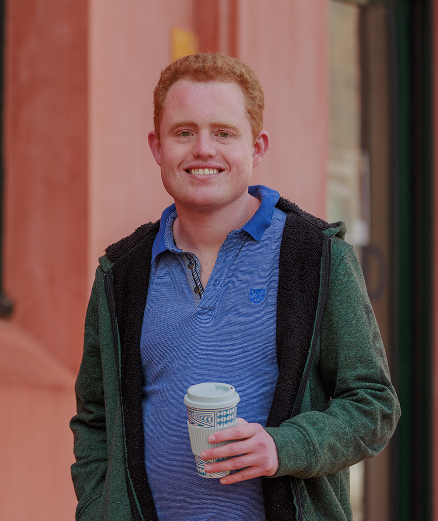 Mitchell stands outside a red office building, holding a takeaway coffee. Mitchell has short red hair and is wearing a bright blue t-shirt underneath a dark green jacket.