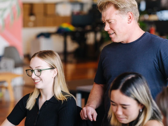 Tianni sits typing at her laptop. She wears green glasses and has long blonde hair. Jonno stands behind her, resting a hand on her swivel chair. He has short blonde hair and wears a navy t-shirt.